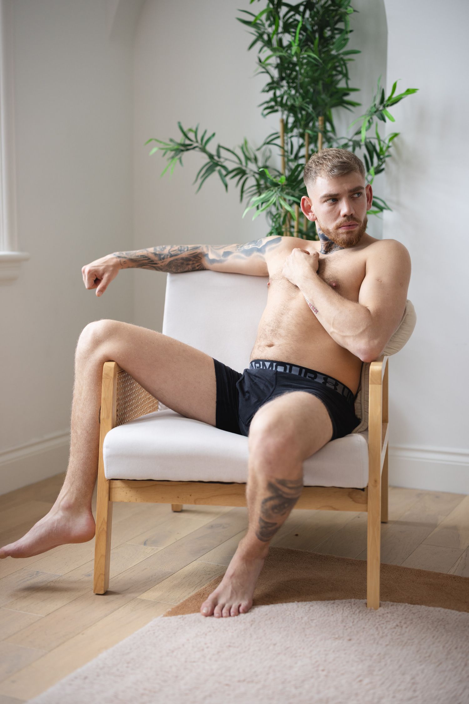Dudeoir photography of a man in black underwear lounging in a wooden chair with a white cushion next to a potted bamboo plant.