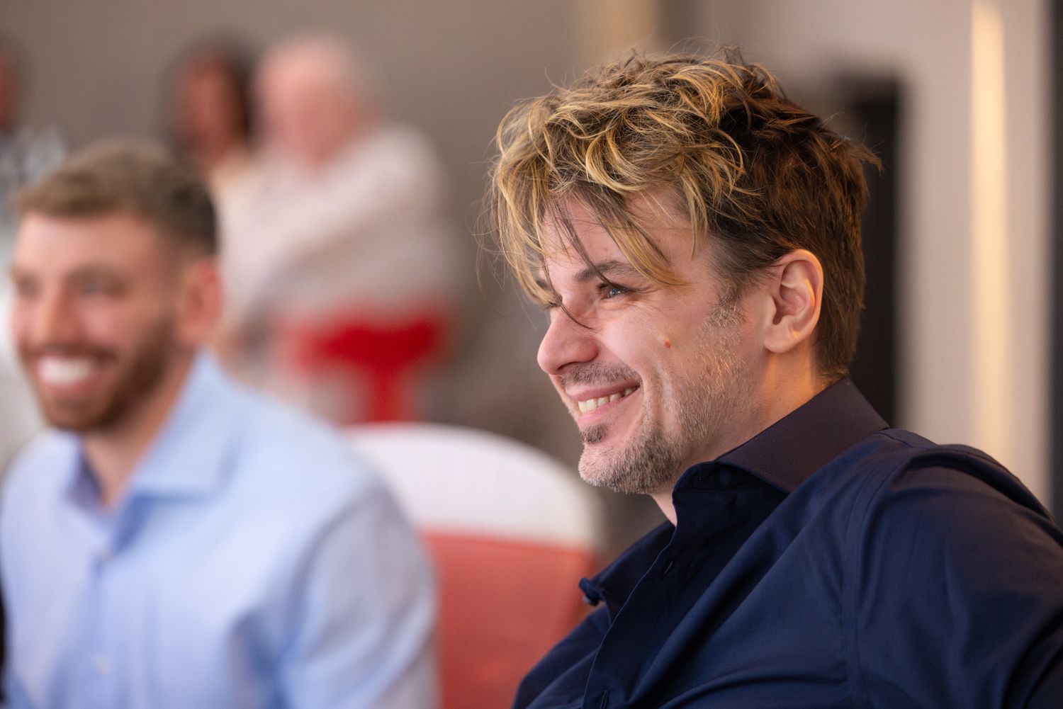 Someone wearing a navy blue collar shirt smiles and looks off to the side while seated indoors.