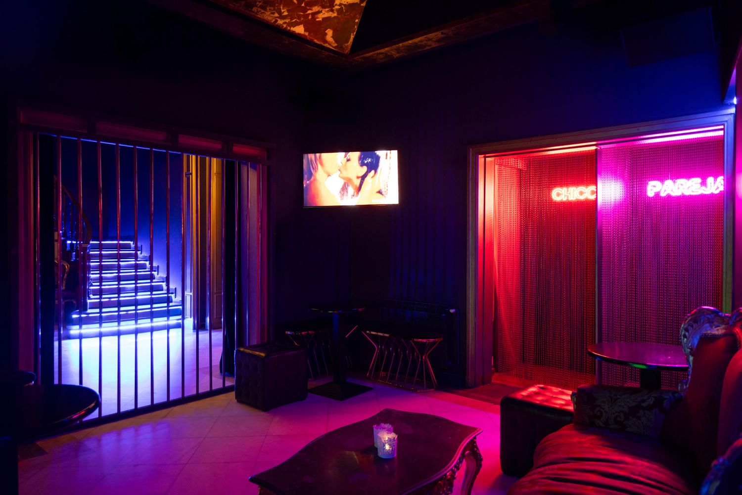 A dimly lit sex club interior with neon pink and blue lighting creating a moody atmospheric ambiance.