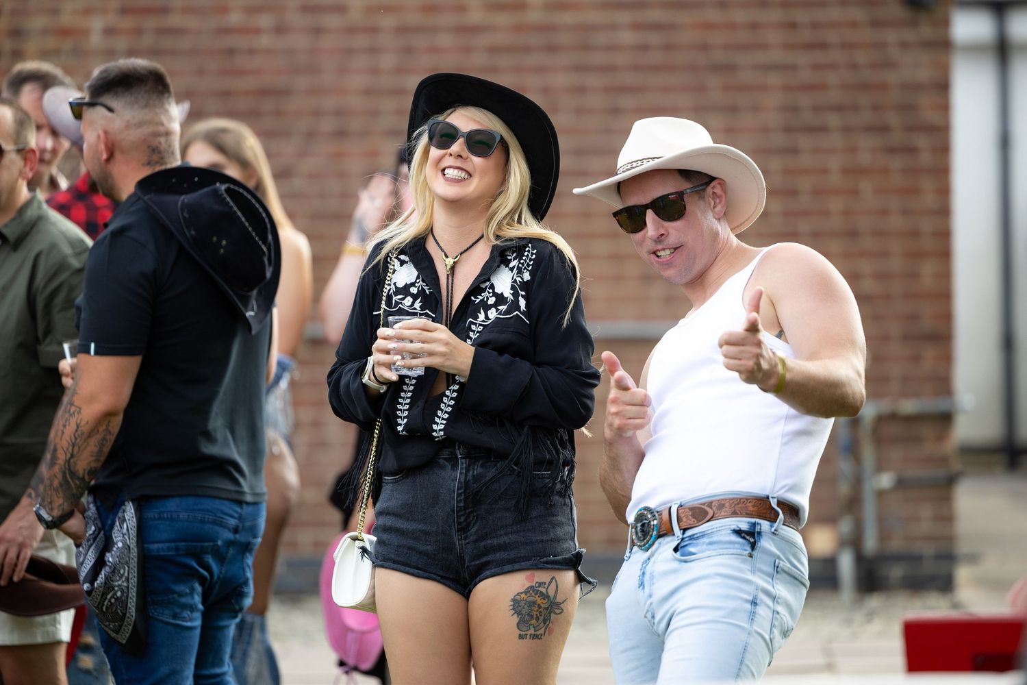 Two smiling people in cowboy hats enjoying an outdoor country music festival, woman in black jacket and denim shorts.