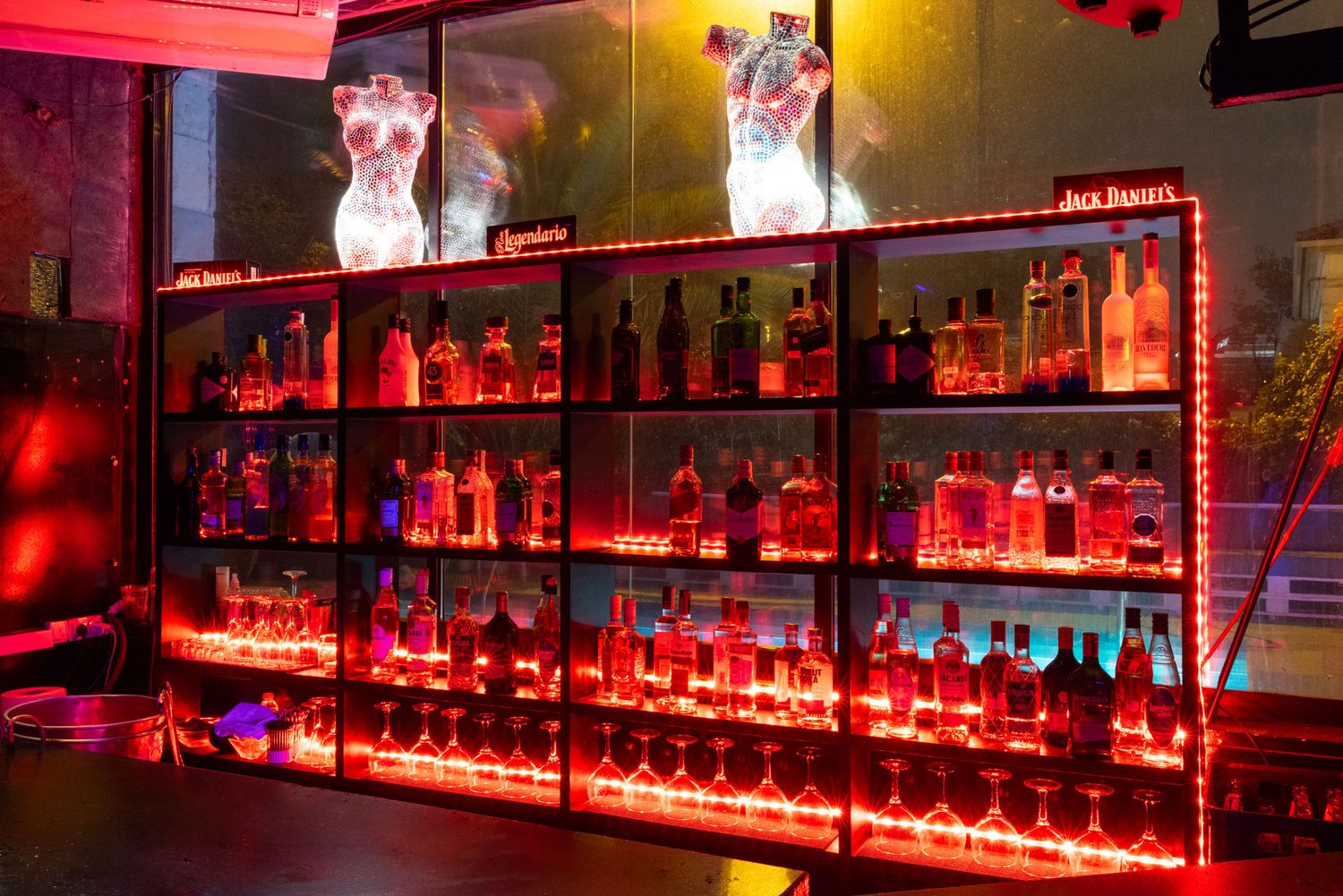 Sex club in Barcelona, a bar shelf illuminated with red lighting displays multiple bottles of alcohol beneath sports images on the wall.