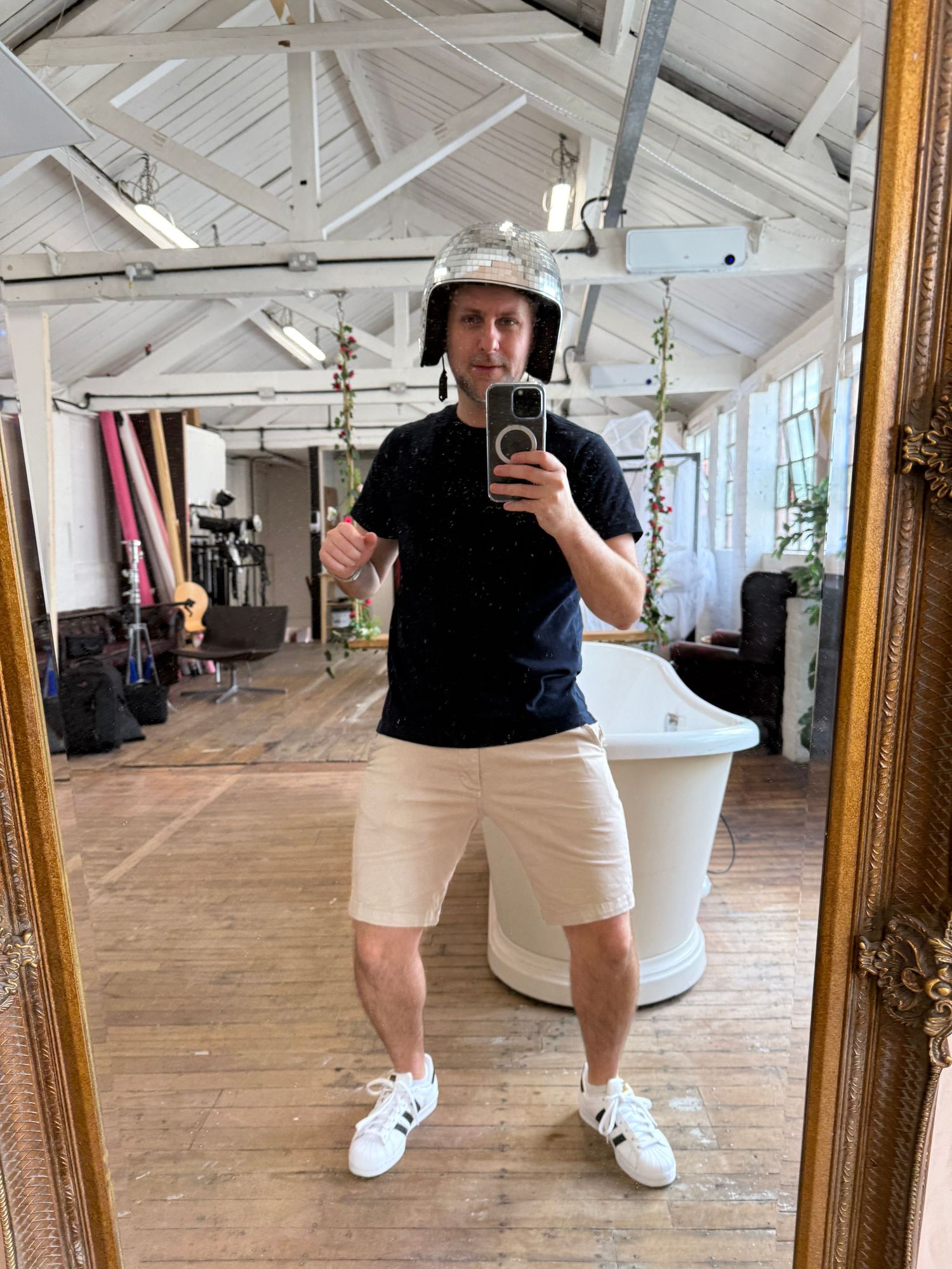 Man in black t-shirt, beige shorts, white Adidas sneakers, and helmet takes mirror selfie in stylish loft space.