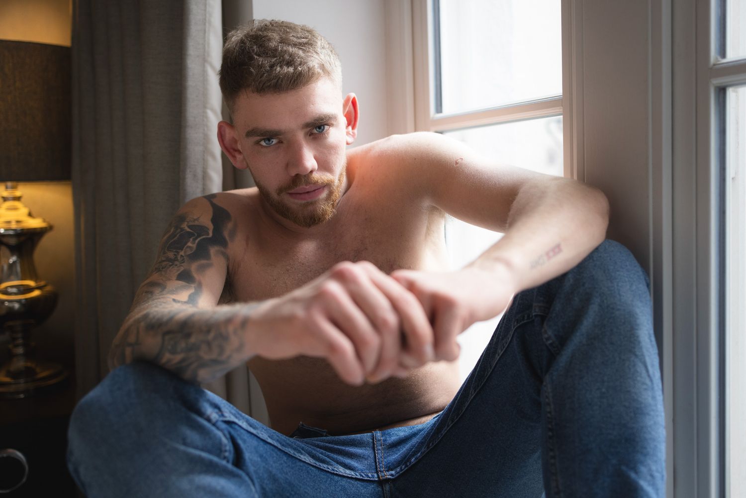 Shirtless man with tattoos sitting near window in casual pose against soft interior lighting and shadows.