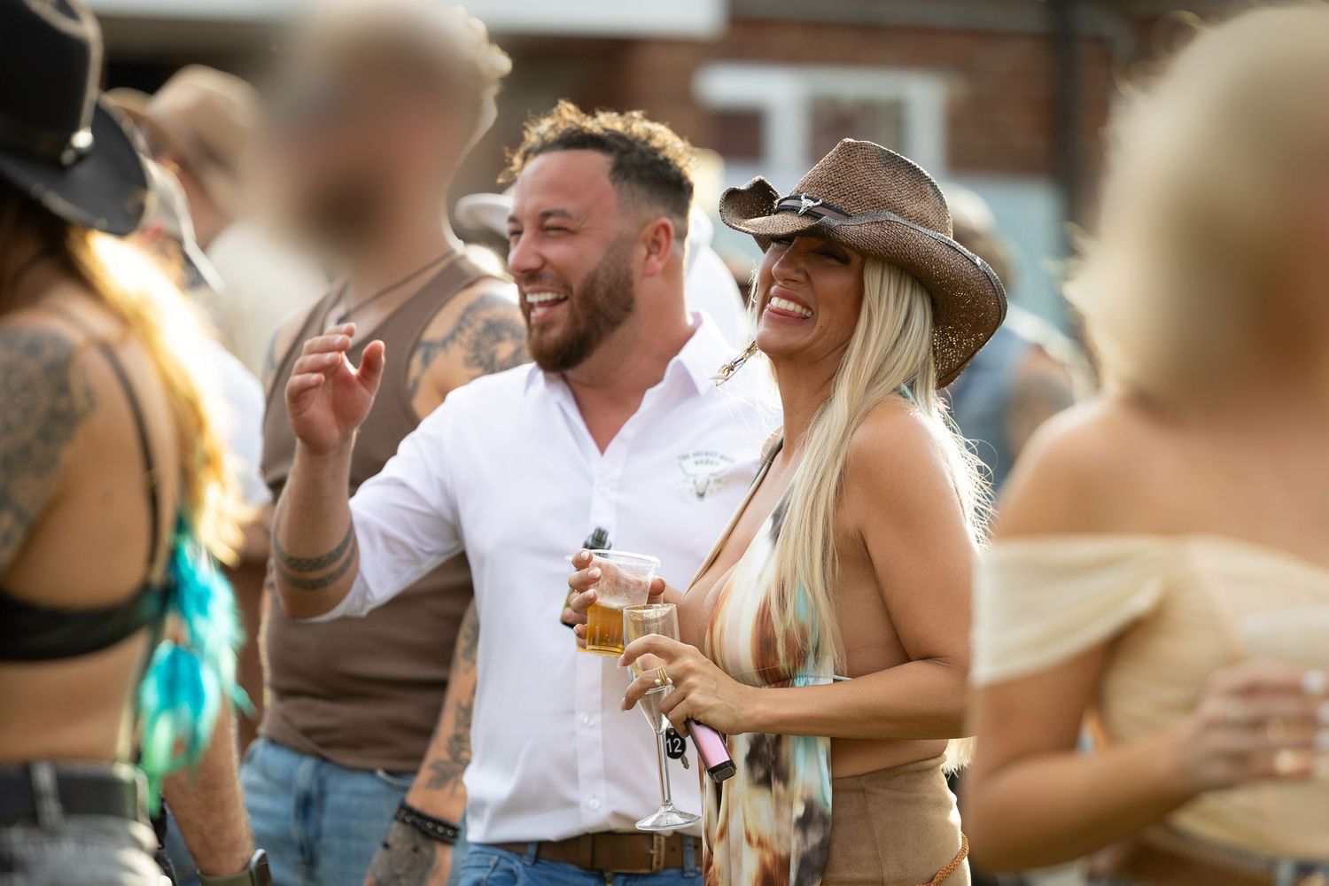 Man in white shirt and woman in cowboy hat laughing together at an outdoor party with drinks.