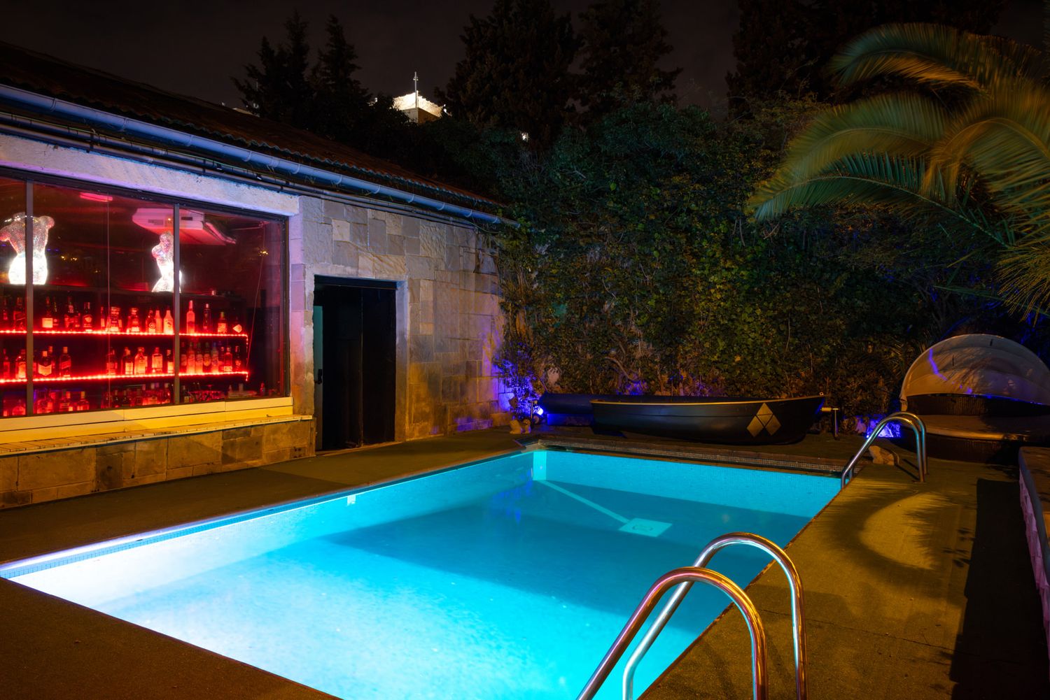 An illuminated swimming pool at an adult club with a glowing blue bar area and palm trees in the background.