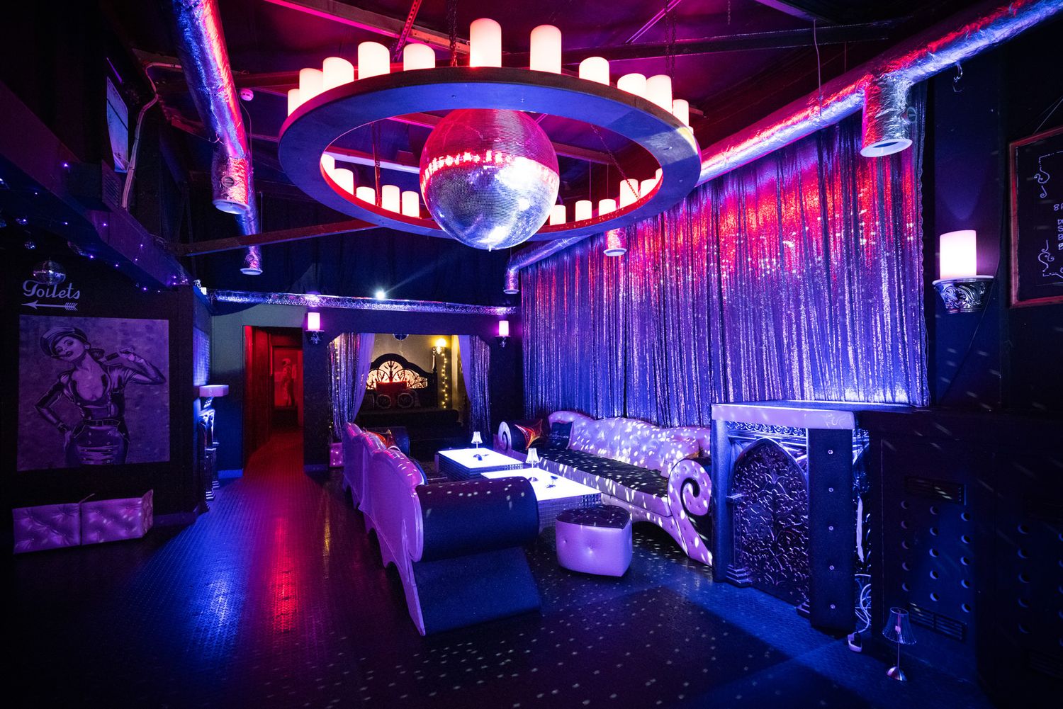 Modern adult club interior with neon lighting, purple and blue accents, and plush seating along mirrored walls.