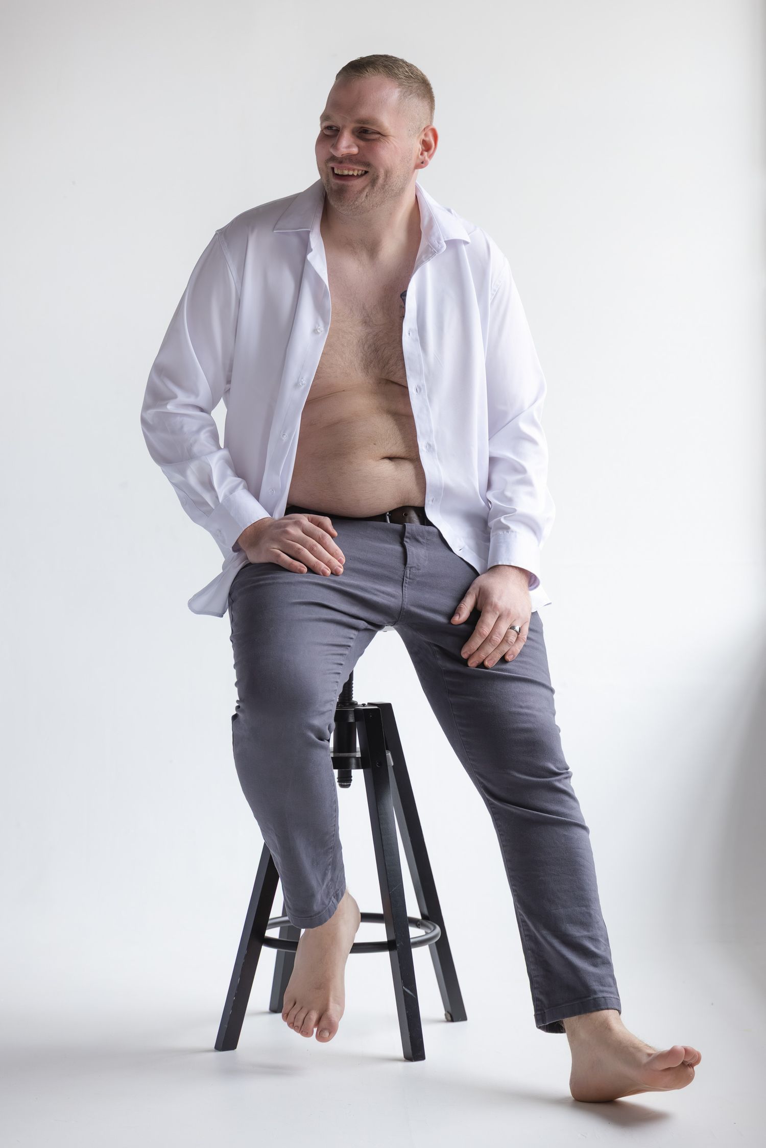 Laughing shirtless man in open white shirt and gray pants sitting barefoot on wooden stool against white background.