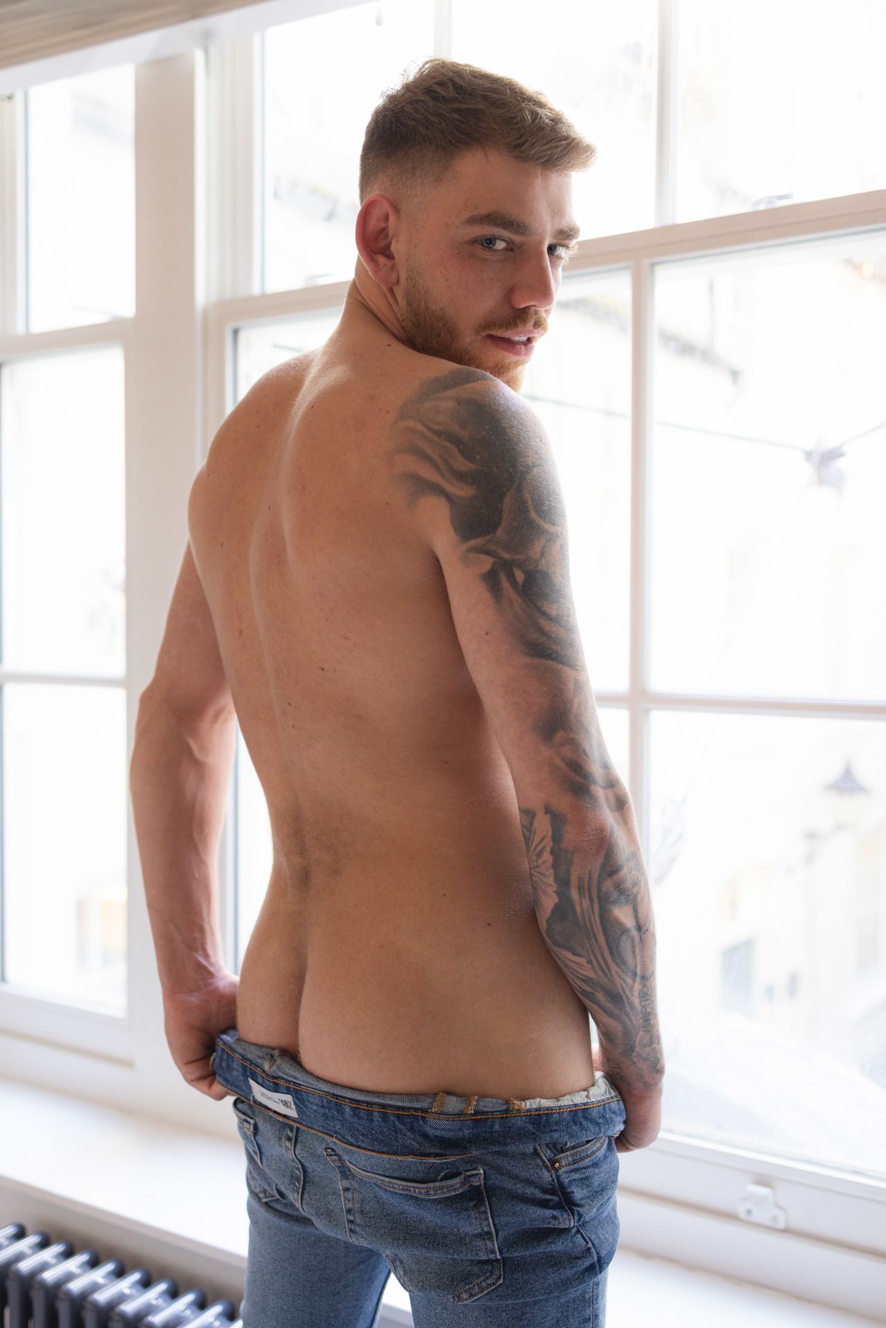 A shirtless person with visible tattoos poses near a window while wearing partially lowered blue jeans.