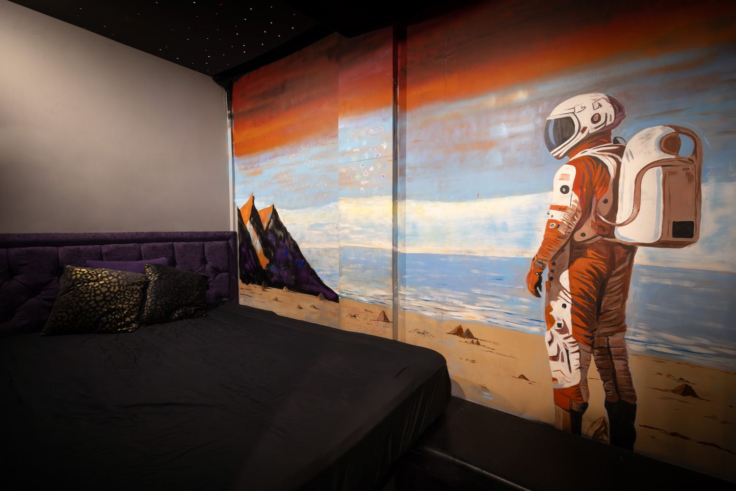 A bed in a swingers club shows a person in orange spacesuit explores scientific equipment against dramatic Mars-like landscape at sunset.