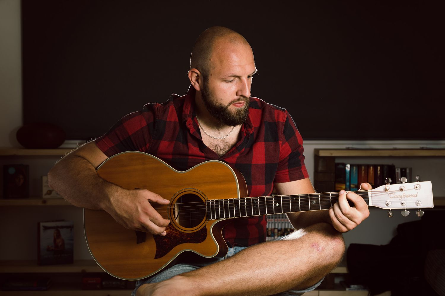 Man wearing red plaid shirt plays an acoustic guitar while sitting indoors in low dramatic lighting.
