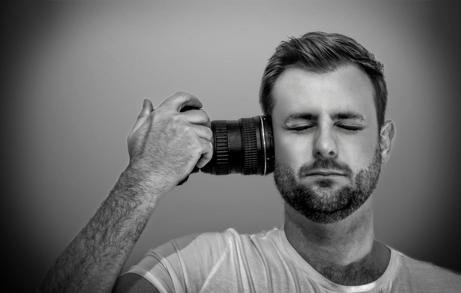 Black and white photo of person holding camera lens to temple with eyes closed wearing white t-shirt.