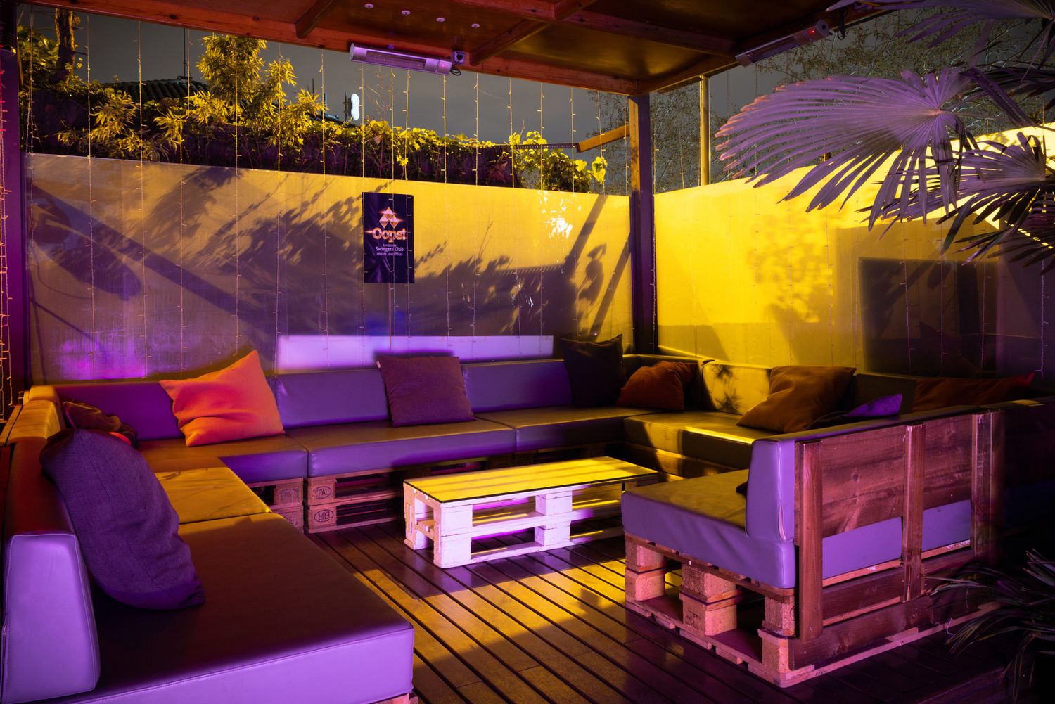 Purple and yellow lights illuminate an outdoor lounge area with white furniture and wooden decking for evening entertainment.