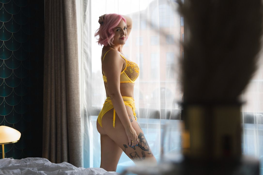 Woman with pink hair wearing yellow lingerie standing by a bright window in a modern hotel room.