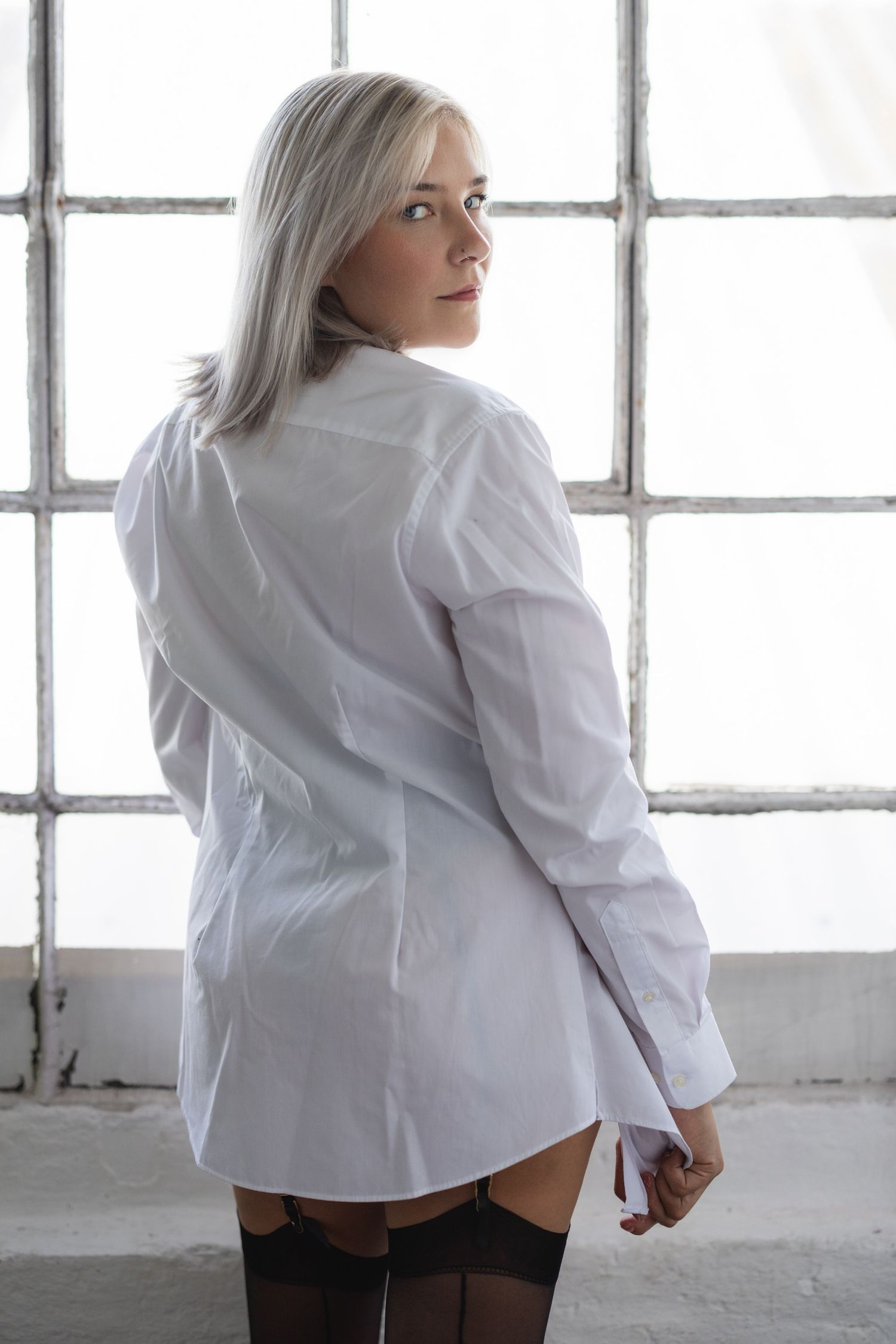 Person in white shirt posing by industrial window in natural light boudoir photoshoot.