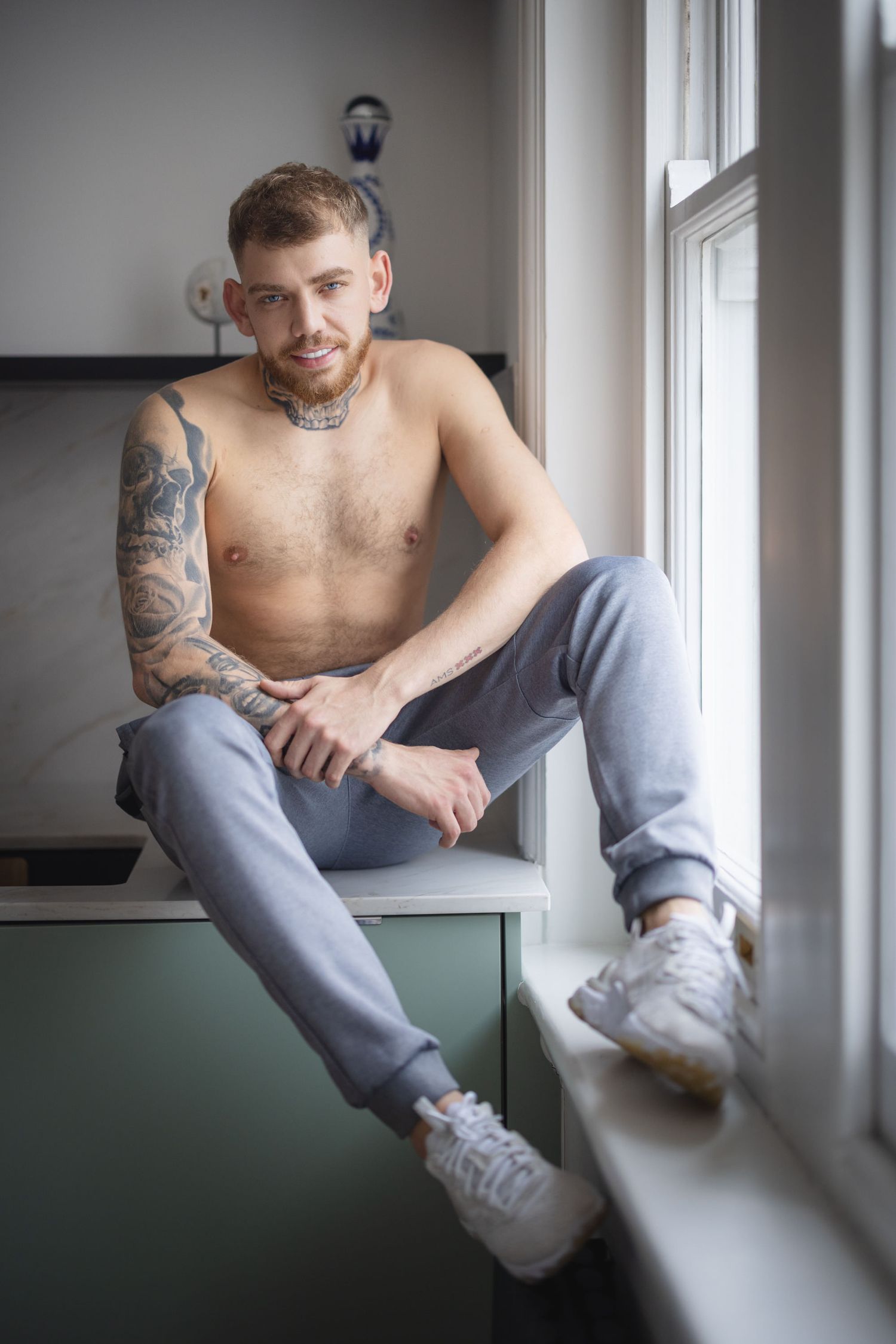 Artistic portrait series showing a shirtless man posing for dudeoir photos in grey sweatpants, lounging by a window in natural light.
