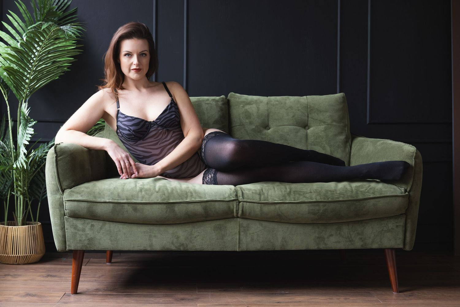 Woman in black bodysuit reclining on green velvet sofa, moody boudoir photography with dark backdrop in Northampton studio
