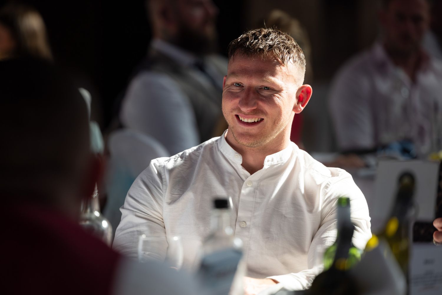 A person in a white button-up shirt sits at a table and smiles warmly in soft lighting during a social gathering.