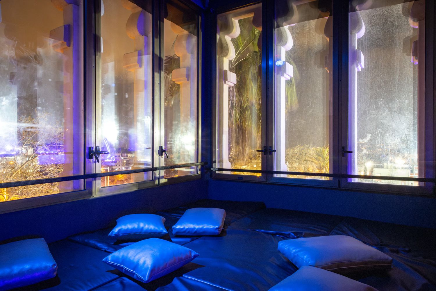 Blue floor cushions arranged at Club Oops in a dim room with large windows showcasing a blurry cityscape at night.