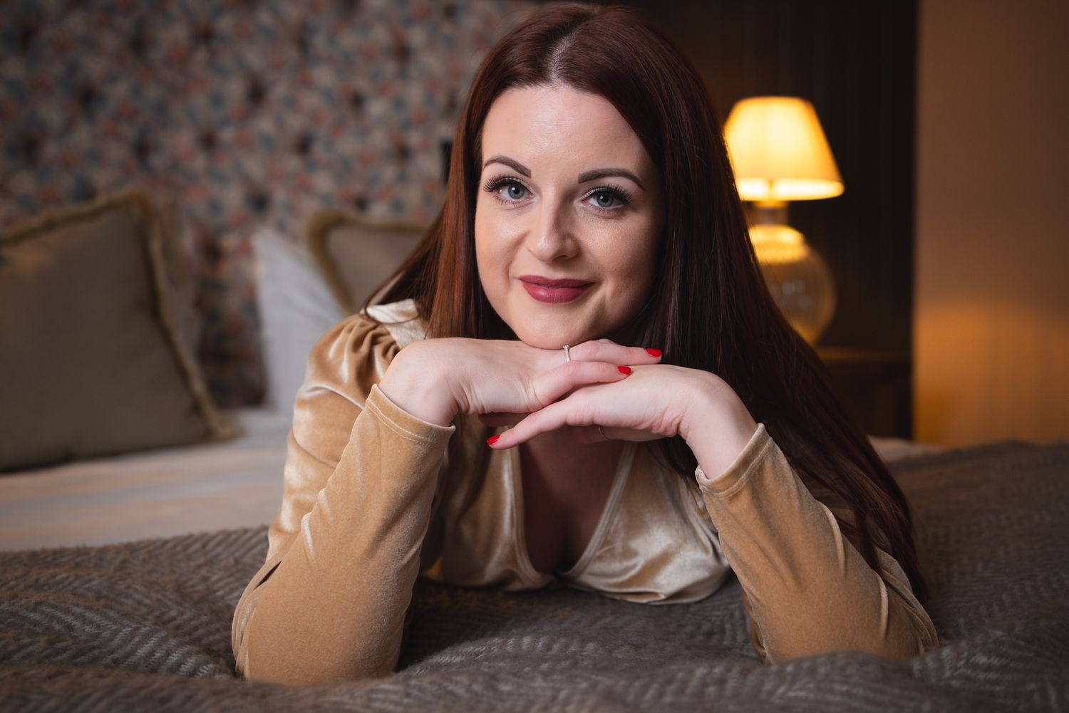 Woman relaxing on bed in cozy bedroom setting with warm lighting and beige sweater.