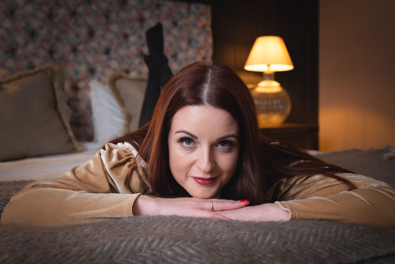 Someone with long dark hair relaxes on a cozy bed in a warmly lit bedroom with a table lamp in the background.