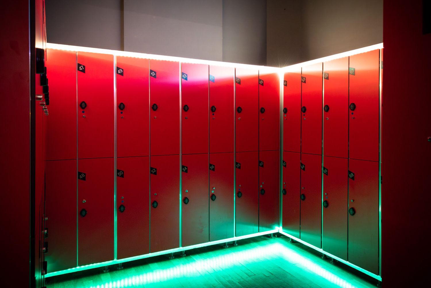 Row of red metal lockers in an adult club locker room with dramatic green and red LED lighting along the ceiling.