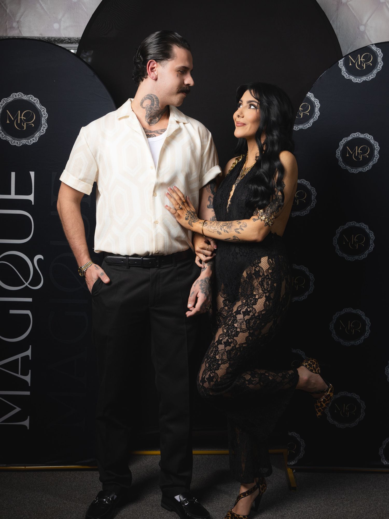 A couple in contrasting outfits poses against a dark backdrop with repeating logo patterns at an adult event.