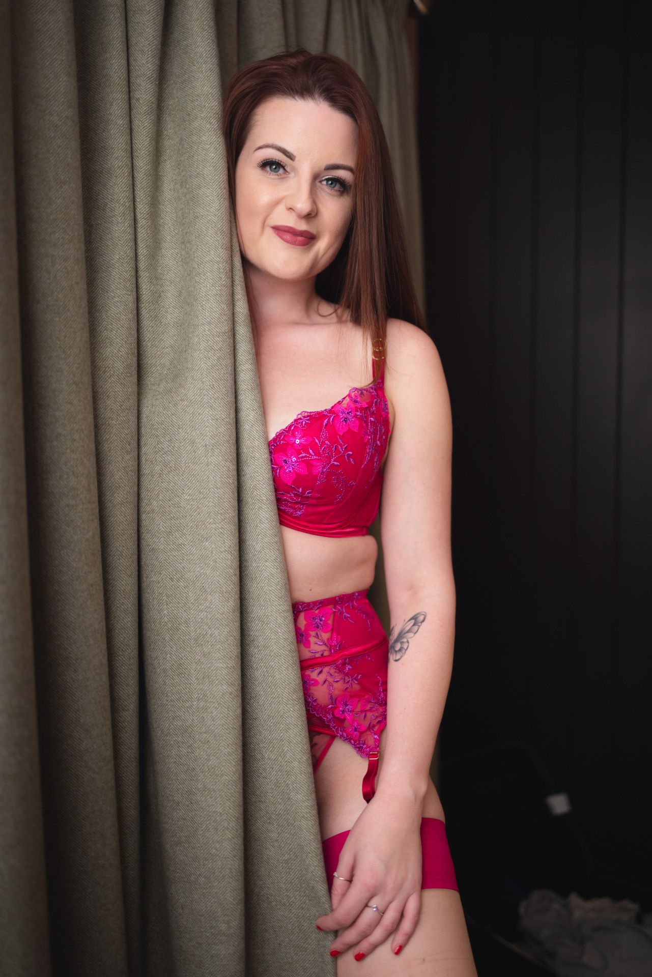 Person wearing vibrant pink lace lingerie set poses beside a beige curtain in an intimate indoor setting.