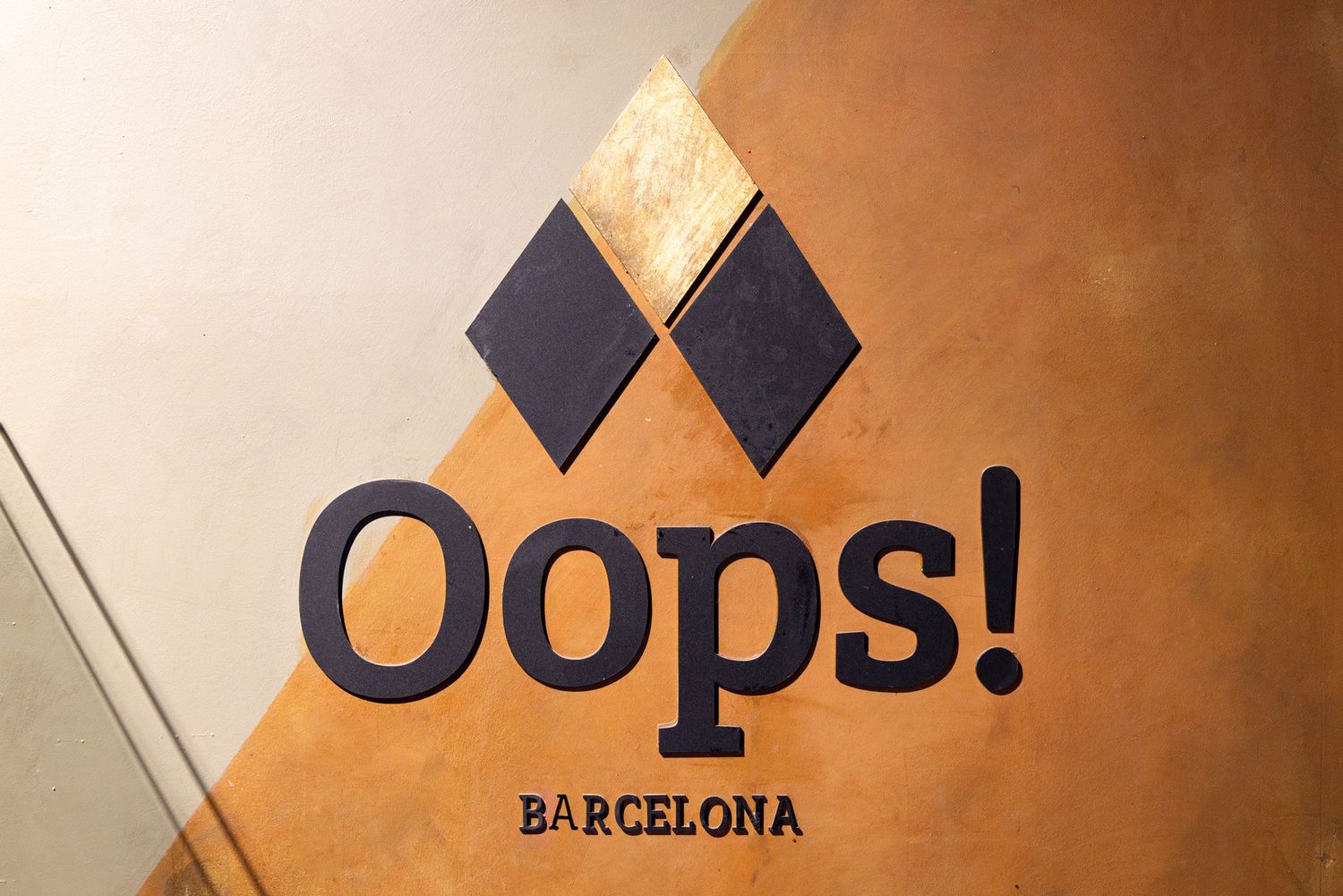 A black and gold logo design for 'Oops! Barcelona' with diamond shapes mounted on a beige wall.