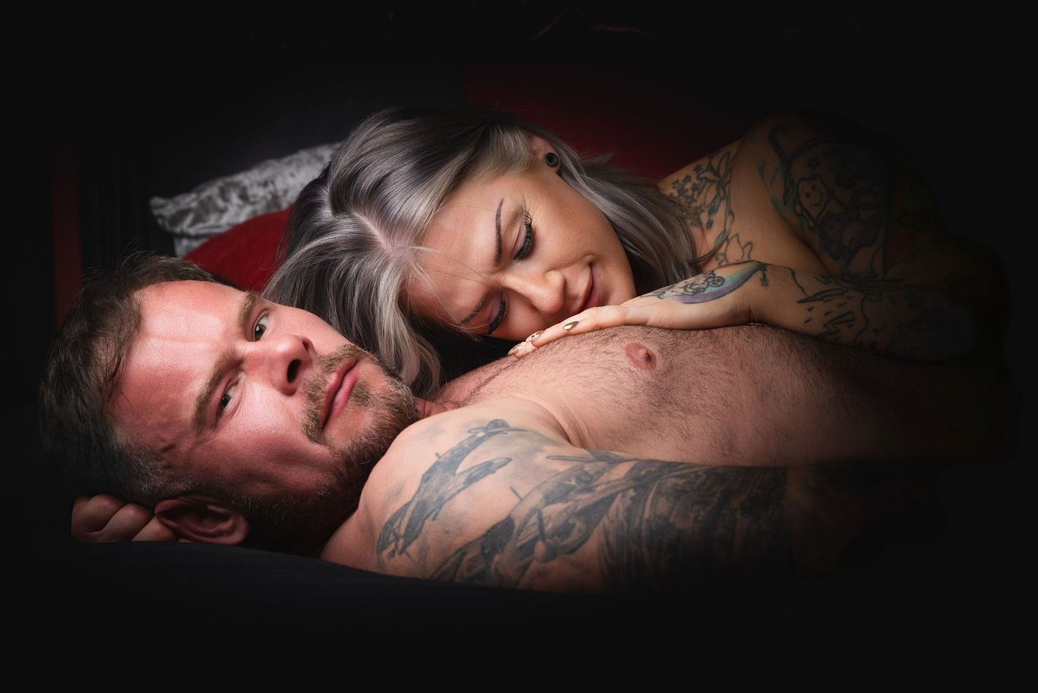 Tattooed couple lying together in bed, woman with gray hair resting her head on man's chest in dim lighting.