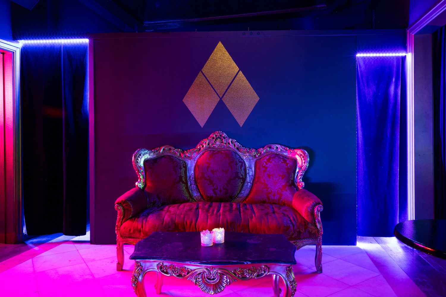 Vintage red velvet sofa illuminated by dramatic pink and blue neon lighting against a dark wall with diamond decor.