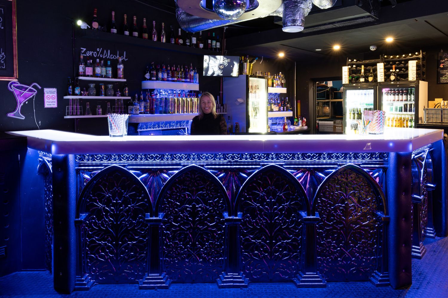 Modern adult club interior with illuminated blue LED bar counters and backlit bottle displays creating ambient lighting.