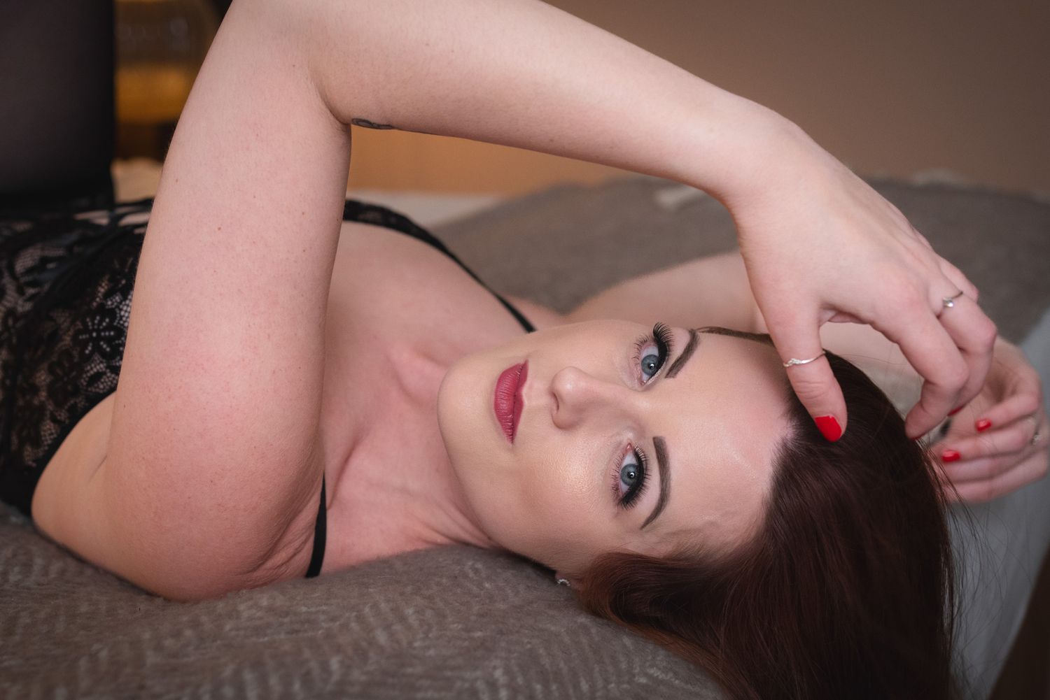 Artistic boudoir portrait shows figure in black lace posing on gray couch with dramatic lighting.