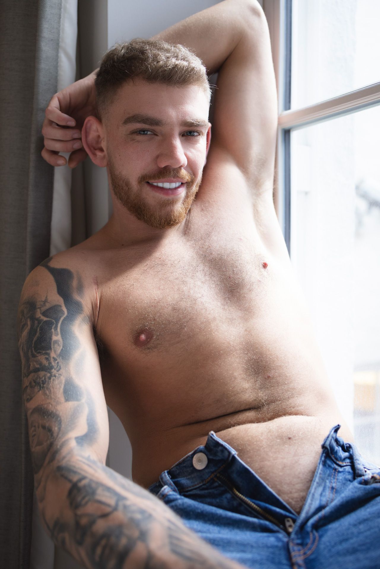 Shirtless person with sleeve tattoos poses next to window wearing blue jeans.