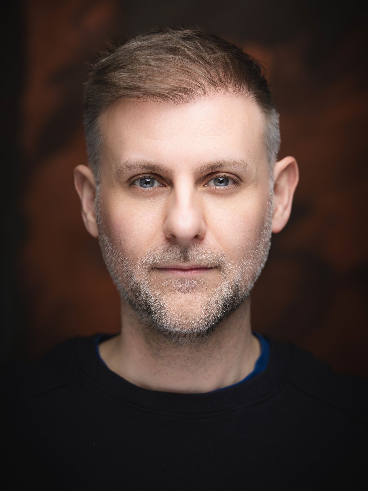 Middle-aged man with blue eyes and salt-and-pepper beard wearing a black top against a dark background.