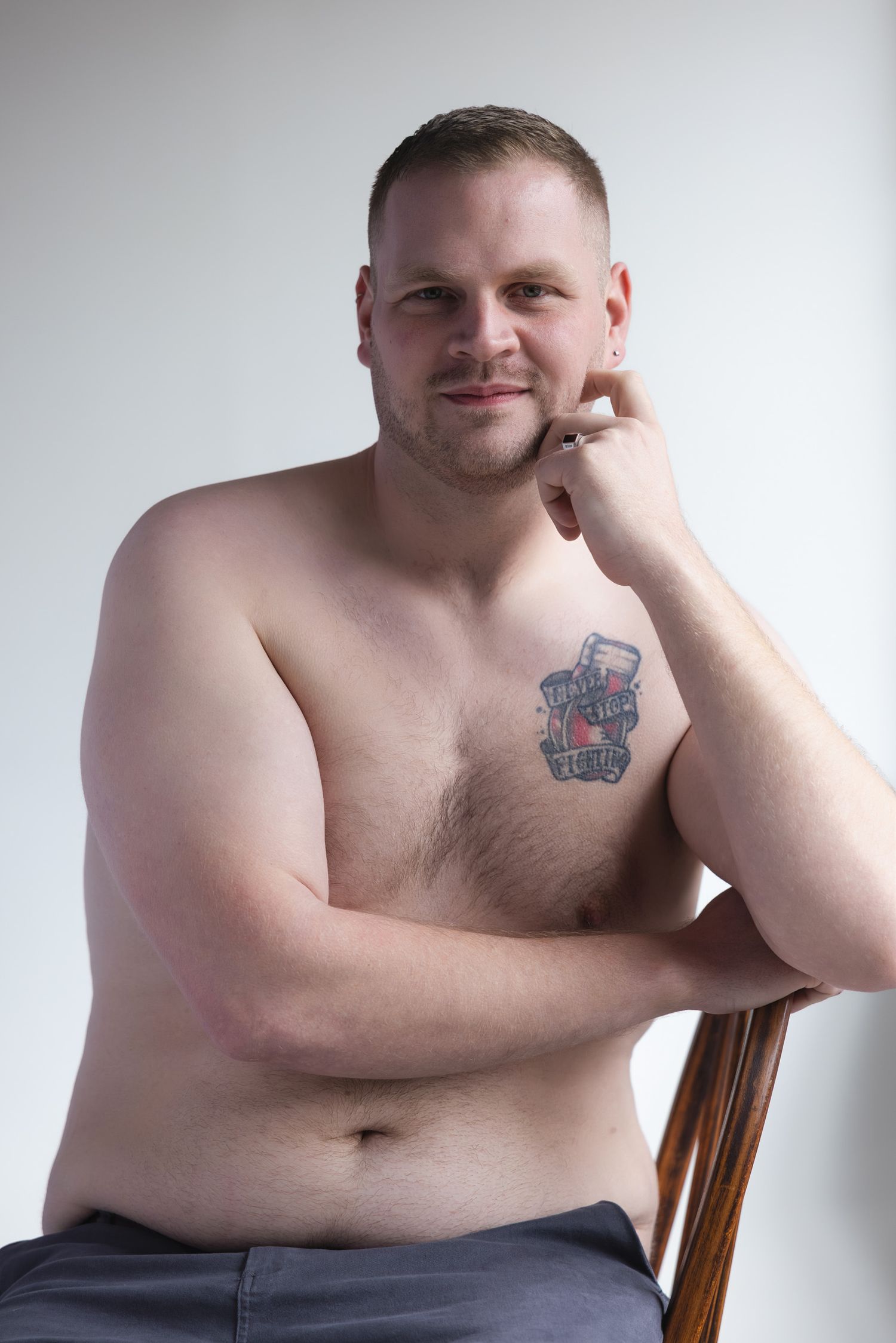 Smiling shirtless plus-size man with colorful chest tattoo posing on chair against white background.