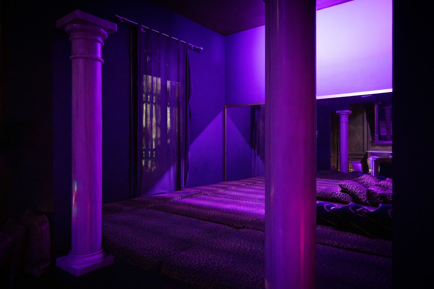 A purple-illuminated bedroom in a sex club creates a moody atmosphere with abstract lighting effects and architectural details.