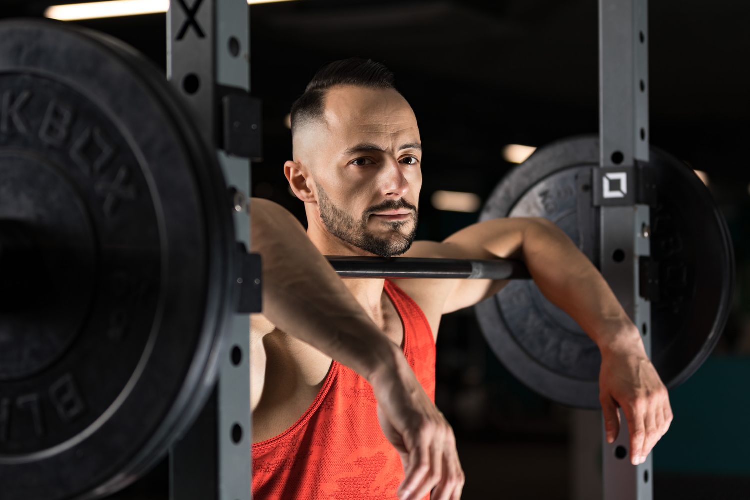 Athletic person in orange tank top taking break during weightlifting session at gym with barbell equipment.