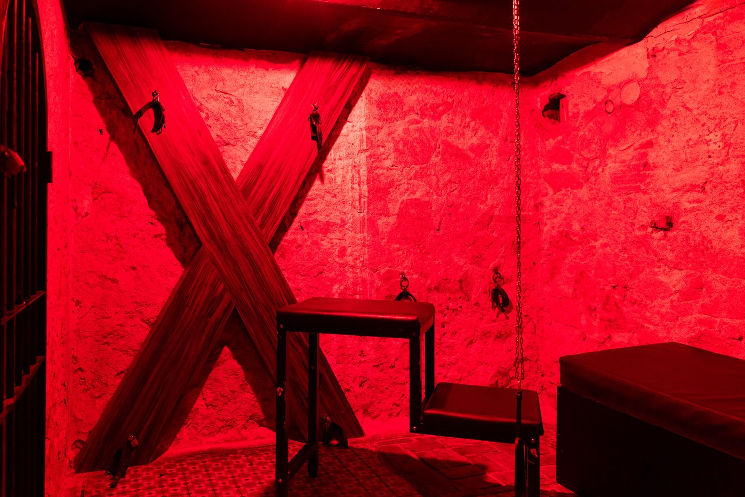 A dark BDSM room illuminated in red light with an X-shaped structure and black angular shapes against the wall.