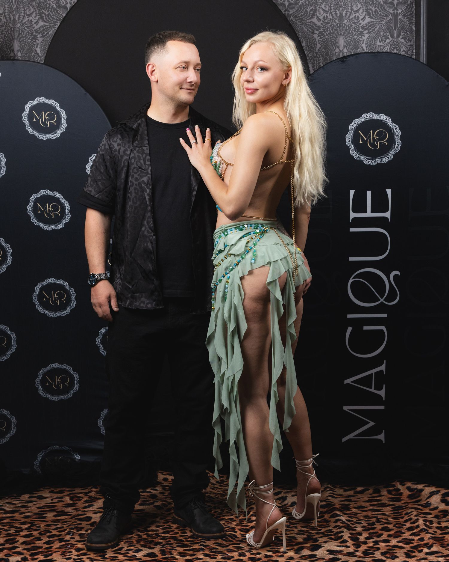 A man and woman pose together at Magique event, she wears a green fringe outfit, he wears black.