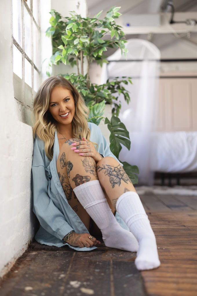 A person in a blue shirt sits on hardwood floor with leg tattoos visible near indoor plants.