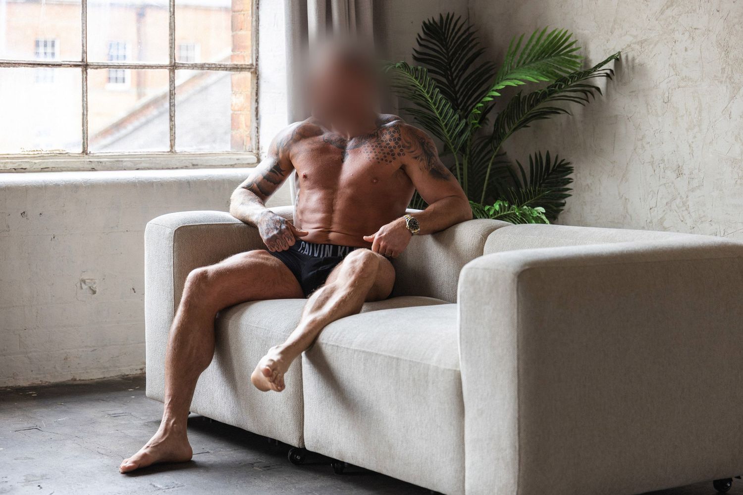 Male boudoir portrait of a muscular tattooed man relaxing on a white sofa in boxer shorts beside a large window and tropical plant.