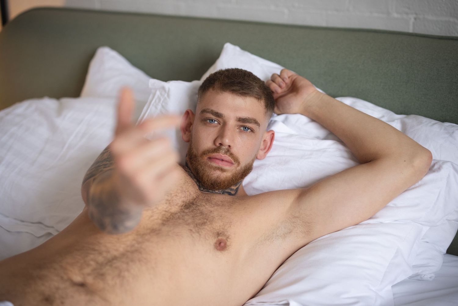 A man lying on white bedding in a relaxed pose with natural lighting, posing for a dudeoir photography session.