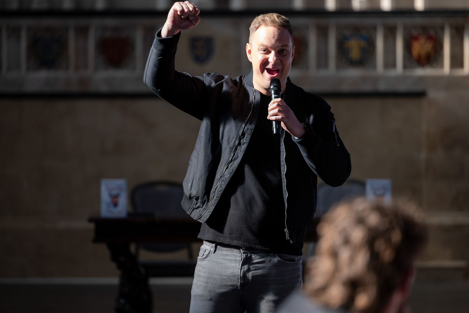A speaker gestures enthusiastically while giving an impassioned speech at an outdoor event with microphone in hand.