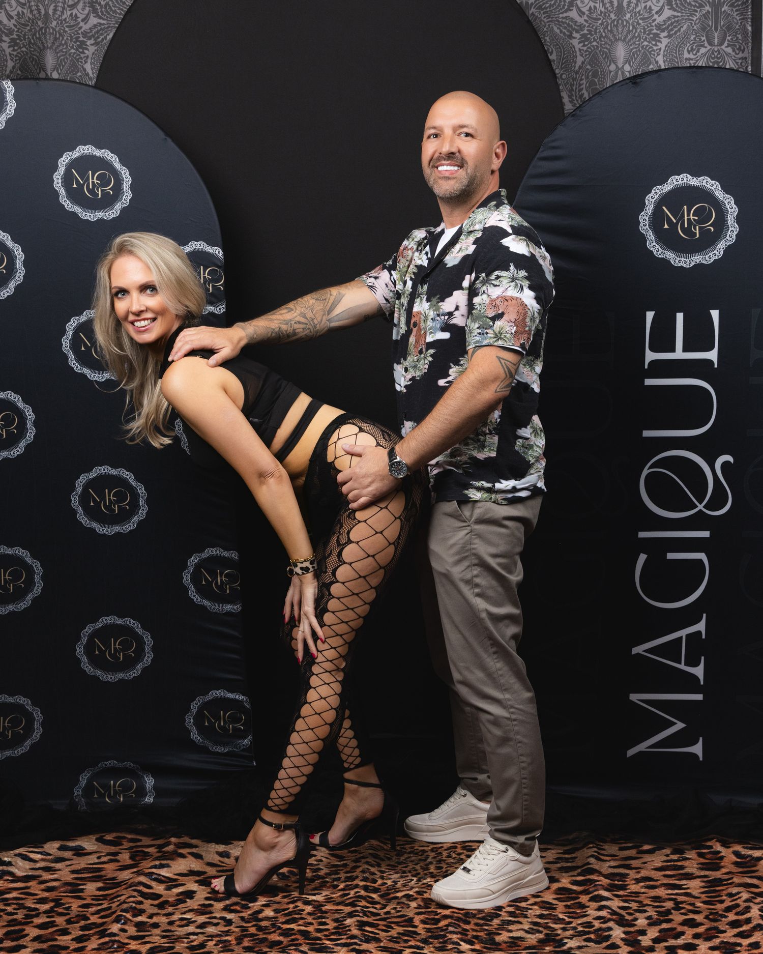 Couple posing at Magique event, woman in fishnet stockings and man in floral shirt on leopard print floor.