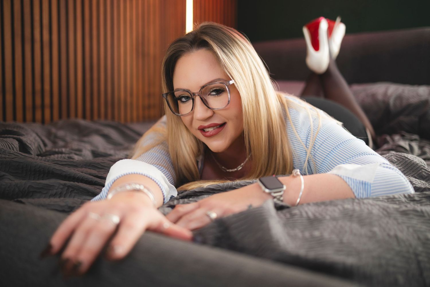 Blonde woman with glasses lying on a bed, smiling at the camera in a cozy indoor setting.
