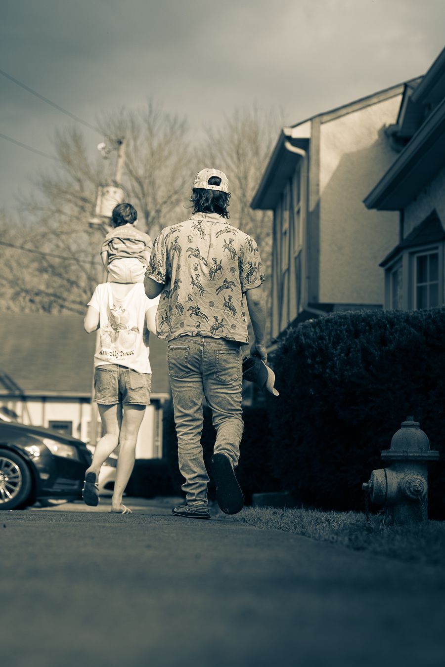 Two people walking down a suburban sidewalk from behind, passing a fire hydrant and parked cars with a child on woman's shoulders.