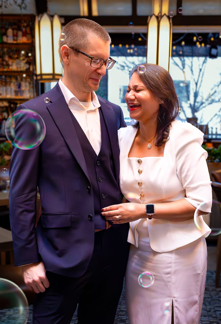 Joyful couple in formal attire laughing together in a bar setting with soap bubbles floating nearby.