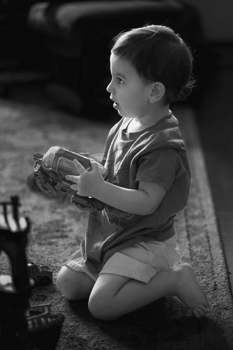 Toddler boy kneeling on carpet holding a toy car, looking away in natural light indoors.