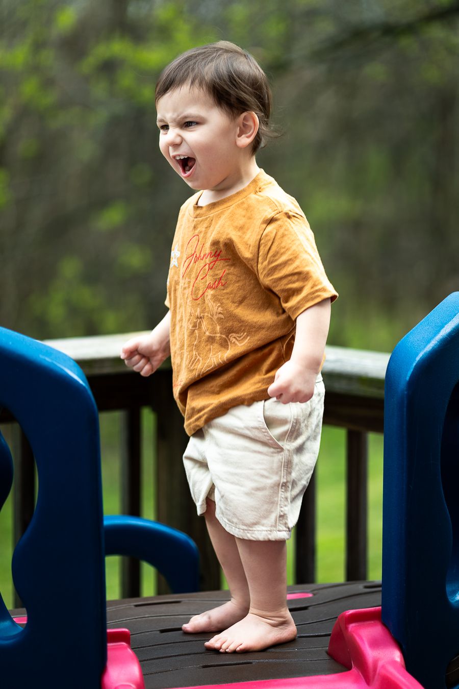 Toddler boy standing on playground toy with mouth open, wearing orange shirt and shorts.