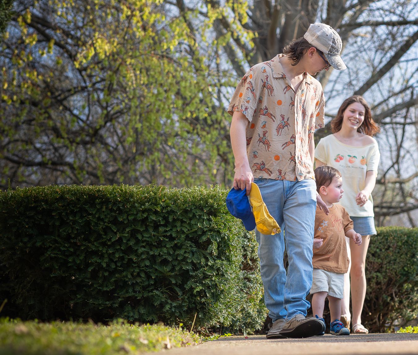A man holding a blue hat walks with a young boy and smiling woman on a sunny outdoor path with young toddler boy